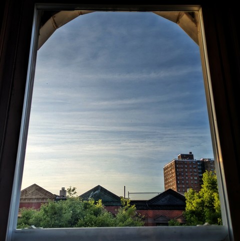 Looking through an arched window an hour after sunrise the blue sky is streaked with horizontal white clouds. Pointed roofs of Harlem brownstones with red brickwork are across the street, and a taller apartment building can be seen in the distance. The green tops of two trees are on the bottom and right.