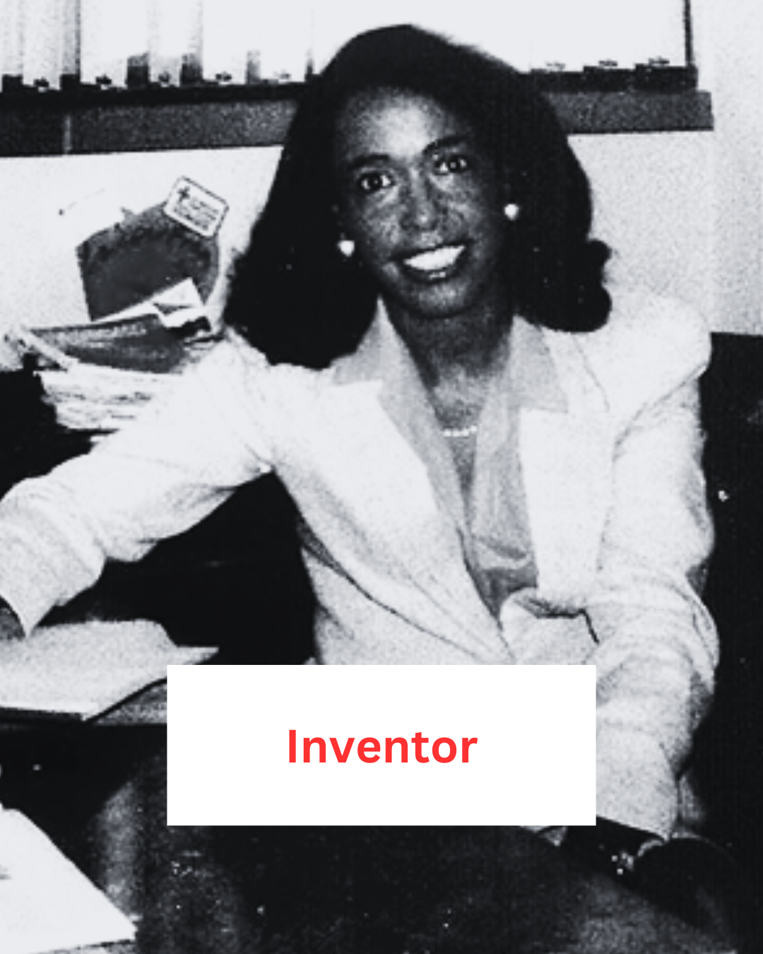 A Black woman in very 1980s clothing, sat behind a desk and smiling. the label reads 'inventor' A Black woman in very 1980s clothing, sat behind a desk and smiling. the label reads 'inventor'