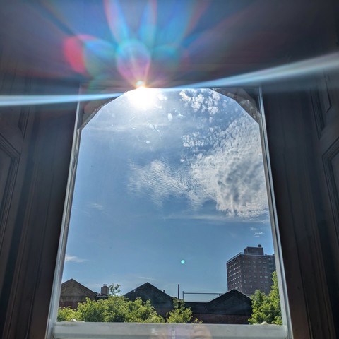 Looking through an arched window three hours after sunrise the sun is high above the buildings, making a dramatic lens flare that bisects the frame diagonally with a curved white line and multi-colored petals above. The sky is light blue with scattered white clouds. Pointed roofs of Harlem brownstones are silhouetted across the street, and a taller apartment building can be seen in the distance. The green tops of two trees are on the bottom and right.