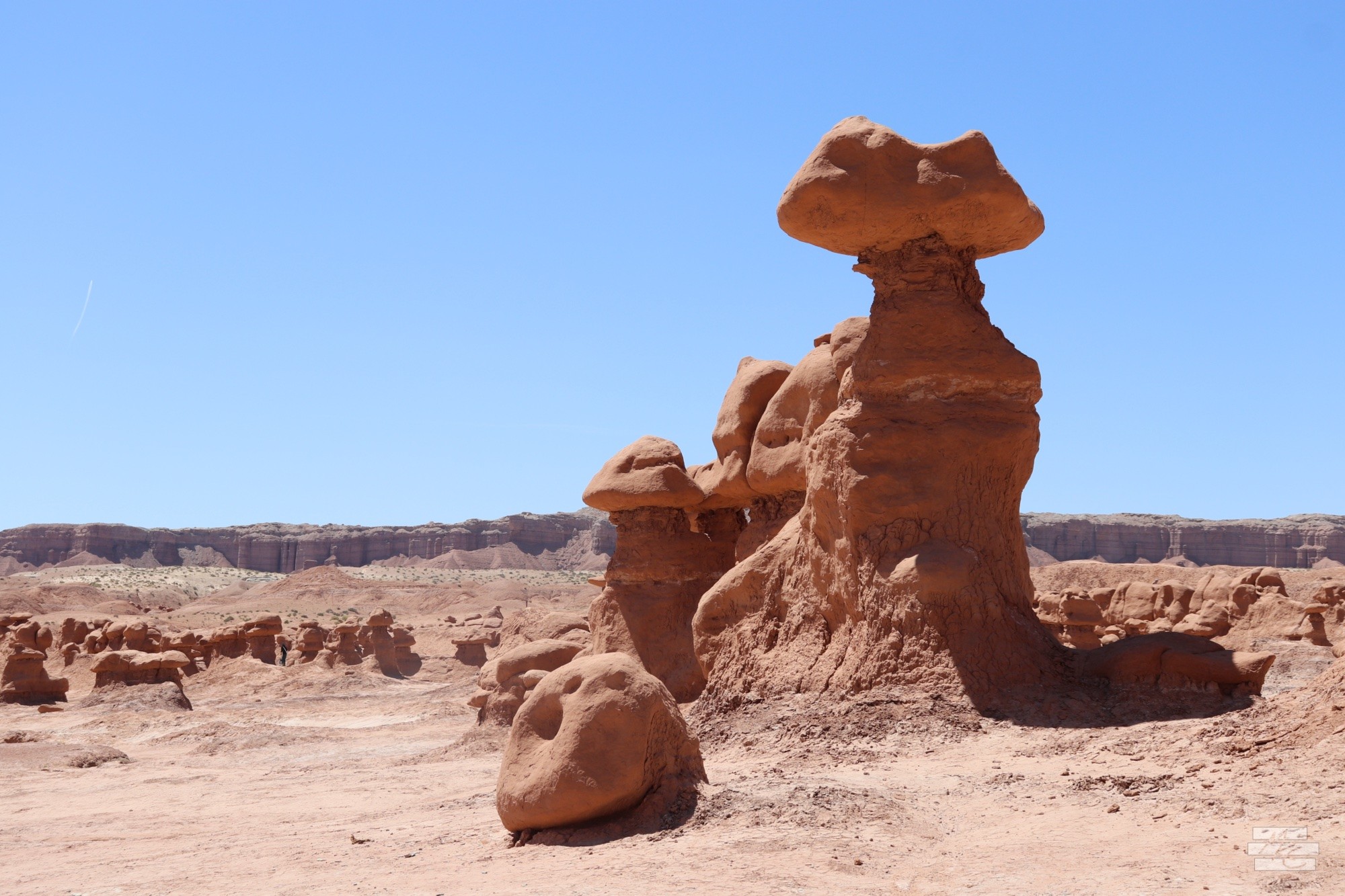 Photograph of a cluster of connected red rock hoodoos with one standout one taller than the rest topped with a large boulder that’s much wider than the column below. Photograph of a cluster of connected red rock hoodoos with one standout one taller than the rest topped with a large boulder that’s much wider than the column below.