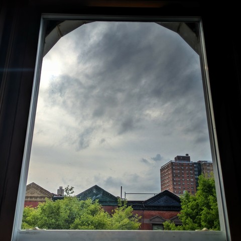Looking through an arched window 2 and 1/2 hours after sunrise the sky is full of complex grey rain clouds. Pointed roofs of Harlem brownstones with red brickwork are across the street, and a taller apartment building can be seen in the distance. The green tops of two trees are on the bottom and right.