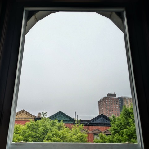 Looking through an arched window ninety minutes after sunrise the sky is full of grey fog. Pointed roofs of Harlem brownstones with red brickwork are across the street, and a taller apartment building can be seen in the distance. The green tops of two trees are on the bottom and right.