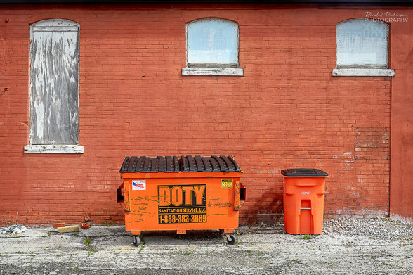 A small orange dumpster on wheels says "Doty Sanitation Service, LLC 1-888-383-3689" on the front.  A smaller orange trash can with a black lid sits next to it.  Both are in front of a brick building with boarded up windows.