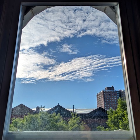 Looking through an arched window two hours after sunrise the bright blue sky is half covered by sinewy white clouds. Pointed roofs of Harlem brownstones are silhouetted across the street, and a taller apartment building can be seen in the distance. The green tops of two trees are on the bottom and right.