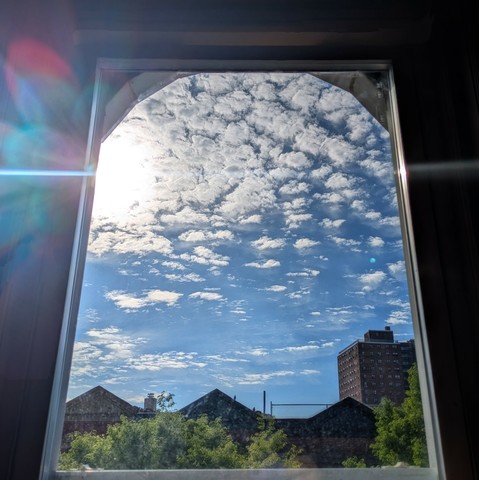 Looking through an arched window two and a half hours after sunrise the sun is high above the buildings on the keft, making a dramatic lens flare that bisects the frame horizontally with a white line and multi-colored petals on the left. The sky is light blue with scattered white clouds. Pointed roofs of Harlem brownstones are silhouetted across the street, and a taller apartment building can be seen in the distance. The green tops of two trees are on the bottom and right.