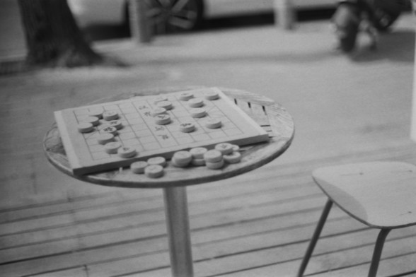 Lucky SHD 400

**English:**
A black and white photograph of a Go board game set up on a small round table outdoors. The board is partially set up with stones placed on it, and there are additional stones in small piles beside the board. The table is situated on a wooden deck, with a stool visible to the right.

**Chinese:**
一张黑白照片，展示了一个围棋棋盘在户外的小圆桌上摆放。棋盘上部分摆放了棋子，旁边有额外的棋子堆放。桌子位于一个木质平台上，右侧可见一个凳子。