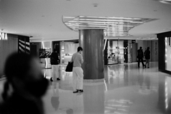 Lucky SHD 400

**English:**
This black and white photograph shows the interior of a modern shopping mall. The scene is characterized by a spacious, well-lit area with a high ceiling. A prominent cylindrical column stands in the center, and the floor is highly reflective, adding to the sleek and polished appearance of the space. Several people are visible, engaged in various activities such as walking and conversing. The mall features various shops, with bright lighting and large display windows…