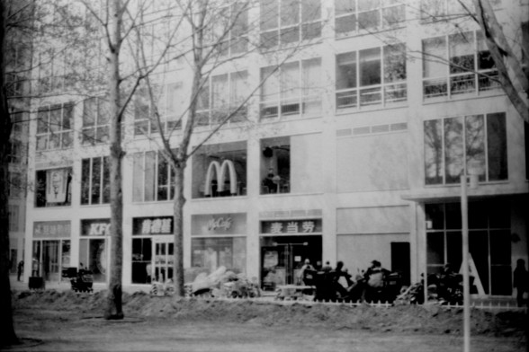 Lucky SHD 400

**English:**
This black and white image depicts the exterior of a commercial building. The building houses a McDonald's restaurant, identifiable by the iconic golden arches logo above the entrance. The facade features large windows, and there are several people visible outside, some of whom appear to be seated on motorcycles. The scene is set in an urban environment, with trees lining the sidewalk and construction materials scattered on the ground, suggesting ongoing development …