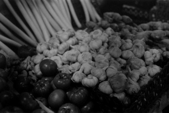 Lucky SHD 400

**English:**
This black and white photograph captures a market scene featuring a variety of fresh produce. In the foreground, there are bunches of garlic with their papery white skins prominently displayed. To the left, there are elongated white radishes, and in the bottom left corner, there are several dark-colored tomatoes. The image highlights the textures and shapes of these vegetables, emphasizing the natural beauty and variety found in a typical market setting.

**Chinese:*…