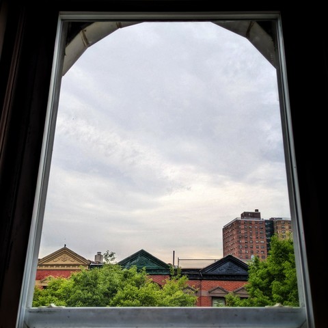 Looking through an arched window eighty-three minutes after sunrise the sky is full of grey clouds, with a subtle tingle of peach at the horizon. Pointed roofs of Harlem brownstones with red brickwork are across the street, and a taller apartment building can be seen in the distance. The green tops of two trees are on the bottom and right.
