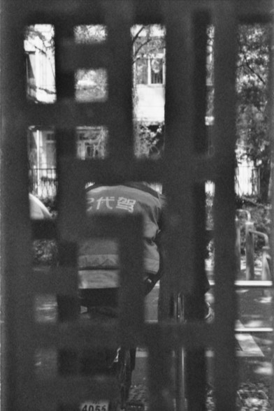 **English Alt Text:**
"A black and white photograph showing a person viewed through a decorative metal fence. The person is wearing a jacket with Chinese characters on the back. The fence has a geometric pattern, and the background includes some greenery and a building."

**Chinese Alt Text:**
"一张黑白照片，通过一个装饰性的金属栅栏可以看到一个人。这个人穿着一件背后有中文字的夹克。栅栏具有几何图案，背景中有一些绿色植物和一栋建筑。"