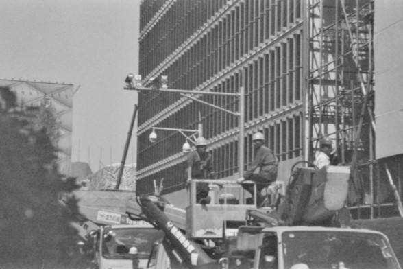 **English Alt Text:**
"A black and white photograph depicting a construction site. Several workers wearing helmets are seen on a raised platform or scaffolding, working on a large structure. The structure appears to be part of a building under construction, with visible beams and structural elements."

**Chinese Alt Text:**
"一张黑白照片，描绘了一个建筑工地。几名戴着安全帽的工人站在一个升高的平台或脚手架上，正在施工一个大型结构。该结构看起来是一栋在建建筑的一部分，可见梁和结构元素。"