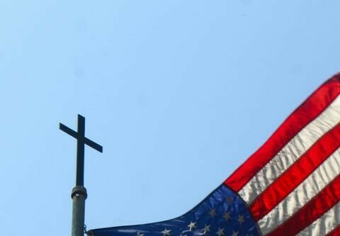 A cross visible at the top of a flagpole over an American flag.