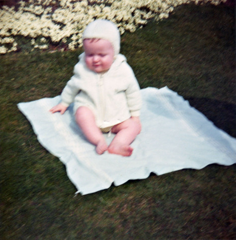 A scan of a colour photo, square, and a little blurry as this is an early 1970s photo and the camera is likely quite a cheap one. We're looking down at a patch of lawn with a border of small white flowers running along the top left corner. In the centre of view there is a square, white blanket, on which there is sitting a very young child, likely only recently able to actually sit at all. The child is wearing a pale green cardigan, done up, and has a cap on his head, also pale green and from the same woollen material.