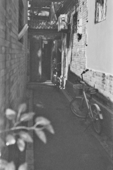 **English:**
A black-and-white photograph of a narrow alleyway with a bicycle parked against the wall. The alley is flanked by brick buildings, and there is a sense of depth created by the perspective. The scene evokes a sense of quiet and simplicity, typical of traditional residential areas.

**Chinese:**
一张狭窄小巷的黑白照片，一辆自行车靠墙停放。小巷两侧是砖房，透视感营造出一种深度。场景传递出一种宁静和简单的感觉，典型的传统住宅区特色。