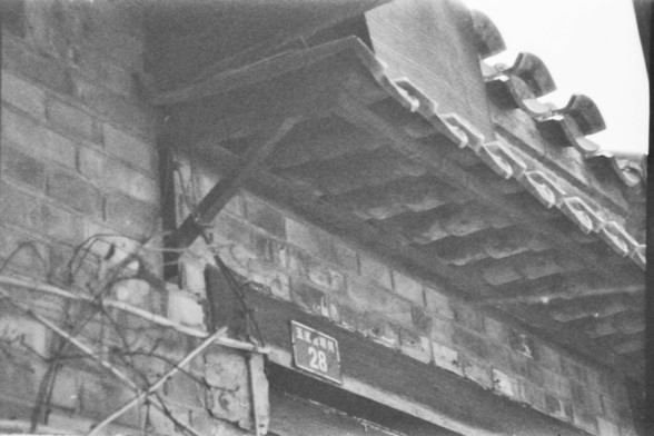 **English:**
A black-and-white photograph of a narrow alleyway between traditional brick buildings. The roof tiles are visible, and there is a sign with the number "28" on the wall. The alley appears to be part of an old residential area, with a rustic and somewhat worn appearance.

**Chinese:**
一张传统砖房之间狭窄小巷的黑白照片。可以看到屋顶的瓦片，墙上有一个写有“28”的标志。这条小巷看起来是老住宅区的一部分，带有乡村风味和略显破旧的外观。