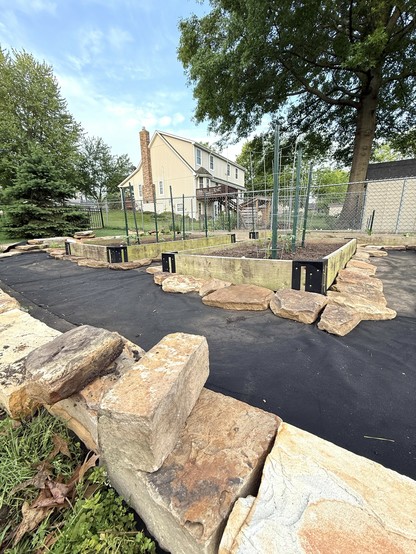 Shot at ground level, the pic shows some flagstone in the foreground, raised beds in the midground and a house and large tree in the background. The morning sky is a light blue with some wisps of clouds. 