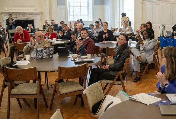 A group of people at tables in a conference room clapping for one person.