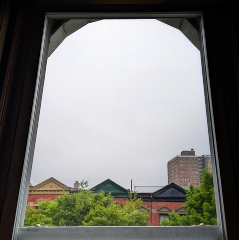 Looking through an arched window an hour after sunrise the sky is full of grey fog. Pointed roofs of Harlem brownstones with red brickwork are across the street, and a taller apartment building can be seen in the distance. The green tops of two trees are on the bottom and right.