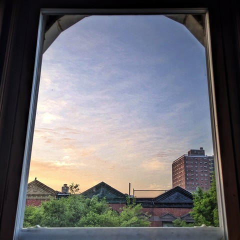 Looking through an arched window twelve minutes after sunrise the light blue sky is full of scattered wispy white clouds, with a peach haze on the bottom left. Pointed roofs of Harlem brownstones with red brickwork are across the street, and a taller apartment building can be seen in the distance. The green tops of two trees are on the bottom and right.