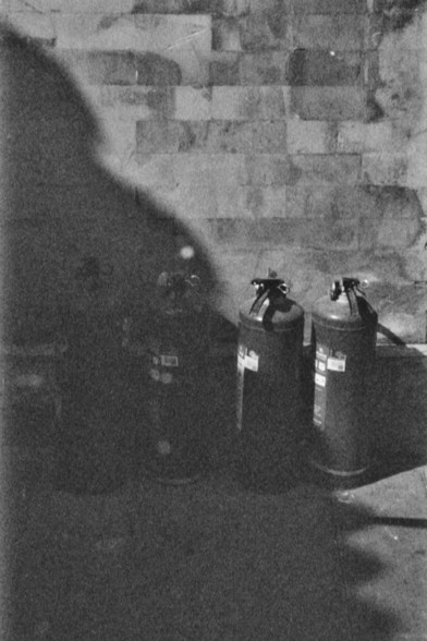 **English:**
A black and white photograph depicting a shadowy scene. The image shows two fire extinguishers placed against a brick wall. The lighting is dim, creating strong shadows and a somewhat mysterious atmosphere. The fire extinguishers are positioned side by side.

**Chinese:**
一张黑白照片，描绘了一个阴影笼罩的场景。照片中有两个灭火器靠着一面砖墙放置。灯光昏暗，形成了强烈的阴影，营造出一种神秘的氛围。两个灭火器并排放置。