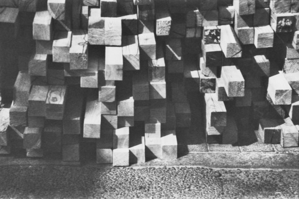 **English:**
A black and white photograph showing a stack of wooden beams. The beams are cut into rectangular shapes and are arranged in a somewhat haphazard manner, creating an abstract pattern of light and shadow. The ground beneath the beams is visible and appears to be made of concrete or asphalt.

**Chinese:**
一张黑白照片，展示了一堆木梁。这些木梁被切割成矩形，并以一种略显杂乱的方式堆放，形成了光影交错的抽象图案。木梁下方的地面清晰可见，看起来是由混凝土或沥青构成的。