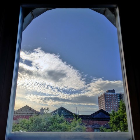 Looking through an arched window ninety minutes after sunrise the bright blue sky is half-full of spiky fluffy white clouds. Pointed roofs of Harlem brownstones with red brickwork are across the street, and a taller apartment building can be seen in the distance. The green tops of two trees are on the bottom and right.