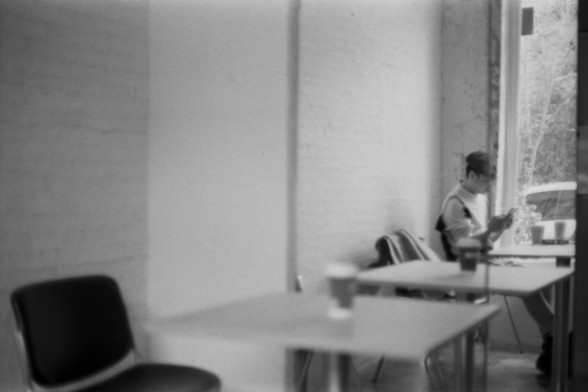 Lucky SHD 400

**English Alt Text:**
"A black and white photograph of an indoor setting. A person is seated at a table near a window, seemingly engaged in an activity, possibly reading or writing. The table has a coffee cup on it. The room has plain walls and another empty table and chair in the foreground."

**Chinese Alt Text:**
"一张黑白照片，展示了一个室内场景。一个人坐在靠近窗户的桌子旁，似乎在进行某项活动，可能是在阅读或写作。桌子上有一个咖啡杯。房间的墙壁很简朴，前景中还有一张空桌子和椅子。"