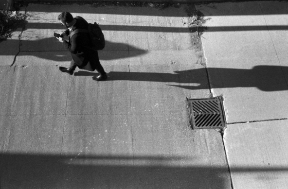 Looking down over a walkway, a man in dark clothes, wearing a backpack, strides past a sewer grate while focussed on his phone. His long shadow stretches behind him. 