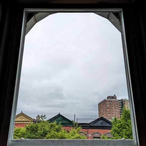 Looking through an arched window an hour after sunrise the sky is full of diffuse grey rain clouds. Pointed roofs of Harlem brownstones with red brickwork are across the street, and a taller apartment building can be seen in the distance. The green tops of two trees are on the bottom and right.
