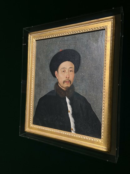 ### English Alt Text:
The image depicts a framed portrait of a man dressed in traditional attire. The man is wearing a black fur hat with a red ornament on top, a black robe with white fur trim, and a white fur stole around his neck. He has a mustache and a goatee. The portrait is encased in an ornate gold frame with intricate detailing, and the background of the painting is a muted gray tone. The portrait is displayed in a modern setting, possibly a museum or gallery, with the frame slightly t…