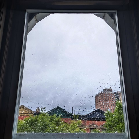 Looking through an arched window an hour after sunrise the sky is full of grey rain clouds and there are raindrops on the glass. Pointed roofs of Harlem brownstones with red brickwork are across the street, and a taller apartment building can be seen in the distance. The green tops of two trees are on the bottom and right.