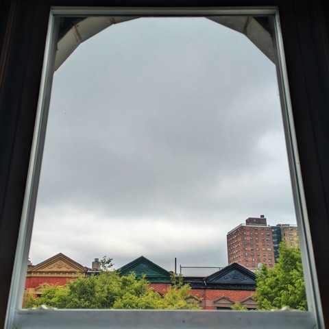 Looking through an arched window four hours after sunrise the sky is full of grey rain clouds. Pointed roofs of Harlem brownstones with red brickwork are across the street, and a taller apartment building can be seen in the distance. The green tops of two trees are on the bottom and right.