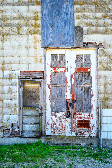 A weathered wood access door is directly next to twin, weathered, white doors in a larger building access opening.  The building is covered in weathered white shingles.  All of the windows in the doors are boarded up.