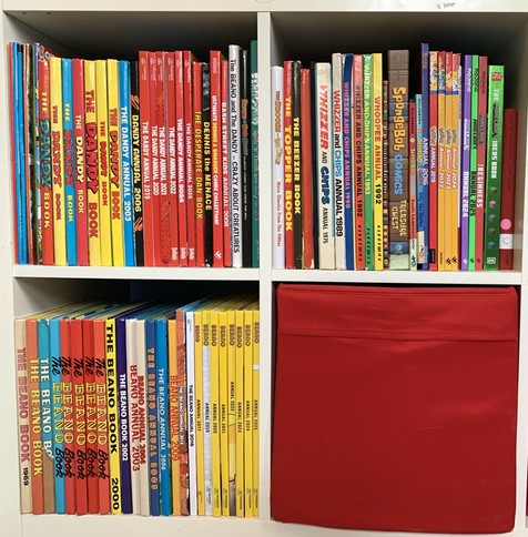 A shelf displaying a variety of colorful comic books and annuals, including titles like "The Beano," "The Dandy," and "SpongeBob Comics." The collection features several editions from different years, arranged mostly by color.