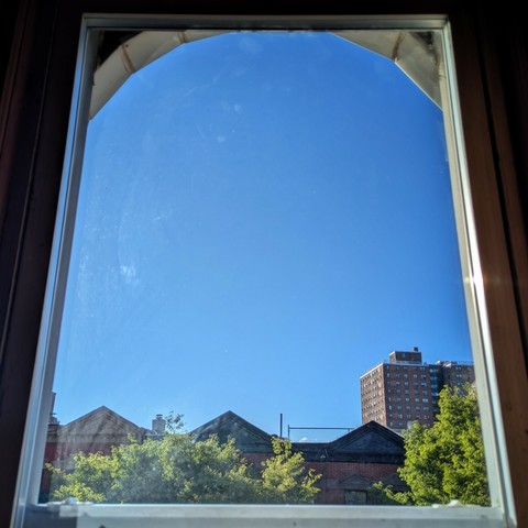 Looking through an arched window an hour and  forty-five minutes after sunrise the sky is deep blue and cloudless. Pointed roofs of Harlem brownstones with red brickwork are across the street, and a taller apartment building can be seen in the distance. The green tops of two trees are on the bottom and right.

