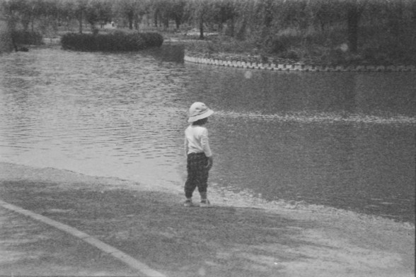 **English:**
A black-and-white photograph of a child standing at the edge of a body of water, possibly a lake or pond. The child is wearing a hat and looking towards the water. The background shows trees and a fence along the water's edge.

**Chinese:**
一个黑白照片，展示一个戴帽子的孩子站在水边，可能是湖泊或池塘。孩子正望向水面。背景中有树木和沿水边的围栏。