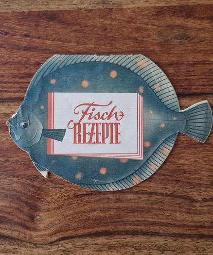 A vintage East German recipe leaflet shaped like a flatfish. The front cover shows the title “Fisch Rezepte” (Fish Recipes) in decorative red script, printed on a stylized fish with orange spots.
