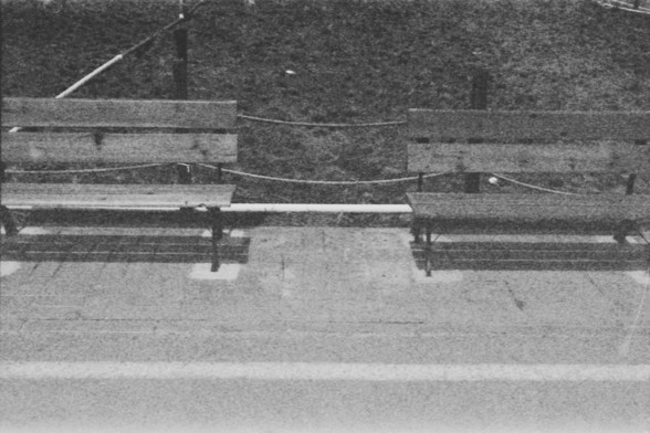 Ilford FP4 @ ISO 100

**English:**
A black and white photograph of two empty wooden benches placed on a paved area. The benches are connected by a metal bar and are situated next to a road. The background is slightly blurred, focusing attention on the benches.

**Chinese:**
一张黑白照片，展示了两张空的木制长椅放置在铺砌区域上。长椅通过金属杆连接，位于道路旁边。背景稍微模糊，将注意力集中在长椅上。