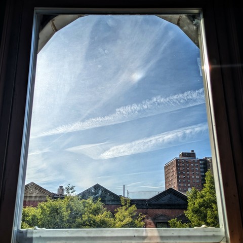 Looking through an arched window two and a half hours after sunrise the blue sky is criss-crossed with large striated white clouds. Pointed roofs of Harlem brownstones are silhouetted across the street, and a taller apartment building can be seen in the distance. The green tops of two trees are on the bottom and right.