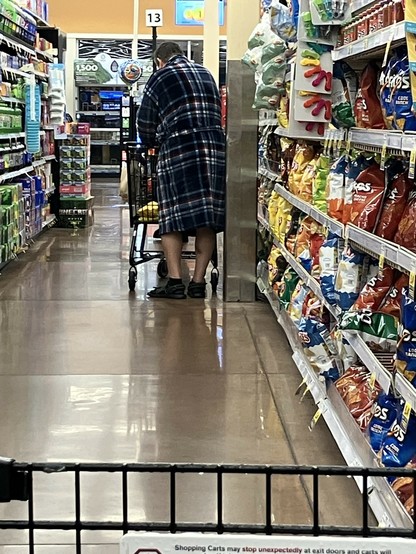 A person wearing a plaid robe and slippers is shopping in a grocery store aisle, standing near a shopping cart filled with items. Snack foods like chips are displayed on the shelves to the right.