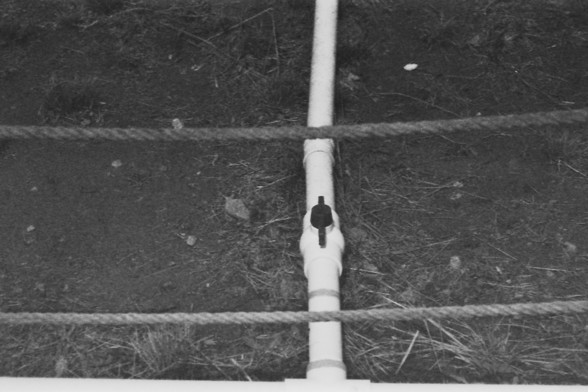 Ilford FP4 @ ISO 100

**English:**
A black and white photograph showing a vertical white pipe with a black connector in the middle. The pipe is positioned in front of a background of horizontal ropes or wires, with a grassy ground visible below.

**Chinese:**
一张黑白照片，展示了一根垂直的白色管道，中间有一个黑色连接器。管道前方的背景是水平的绳索或电线，下方可见草地。