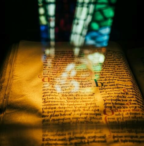 A view of a bible page in old hand written text. In the foreground is a reflection of a stained glass window.