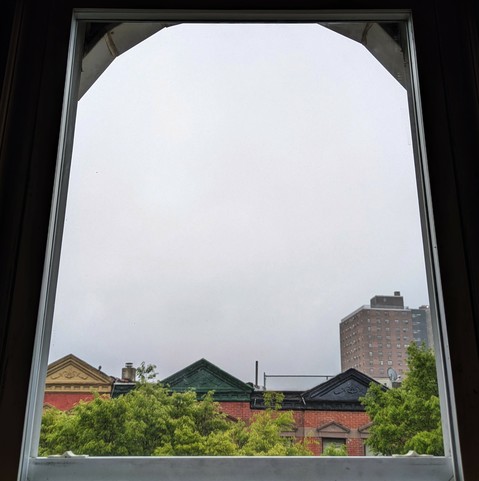 Looking through an arched window two hours after sunrise the sky is full of grey fog. Pointed roofs of Harlem brownstones with red brickwork are across the street, and a taller apartment building can be seen in the distance. The green tops of two trees are on the bottom and right.