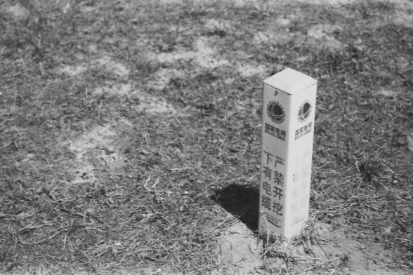 Ilford FP4 @ ISO 80

**English:**
This black and white image shows an electrical warning post situated in a grassy area. The post is rectangular and has text and symbols indicating the presence of underground cables. The grass around the post appears dry and slightly overgrown, suggesting a rural or less-maintained area. The warning post serves as a caution to prevent any accidental damage to the underground utilities.

**Chinese:**
这张黑白照片展示了一个位于草地上的电力警示柱。警示柱呈长方形，上面有文字和符号，指示地下有电缆。柱子周围的草看起来干燥且略显…