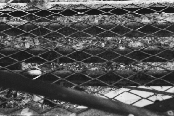 Ilford FP4 @ ISO 80

**English:**
This black and white photograph captures a scene dominated by a chain-link fence in the foreground. The fence creates a sense of separation and barrier. Behind the fence, the image reveals a somewhat cluttered and indistinct background, which may include various objects or debris. The overall mood of the image is somber and reflective, enhanced by the monochromatic tones. The image may evoke themes of confinement, isolation, or abandonment.

**Chinese:**
这张黑白照片…