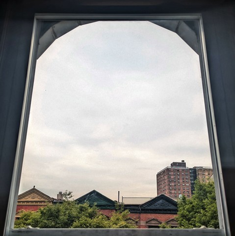 Looking through an arched window four hours after sunrise the sky is full of light grey clouds. Pointed roofs of Harlem brownstones with red brickwork are across the street, and a taller apartment building can be seen in the distance. The green tops of two trees are on the bottom and right.