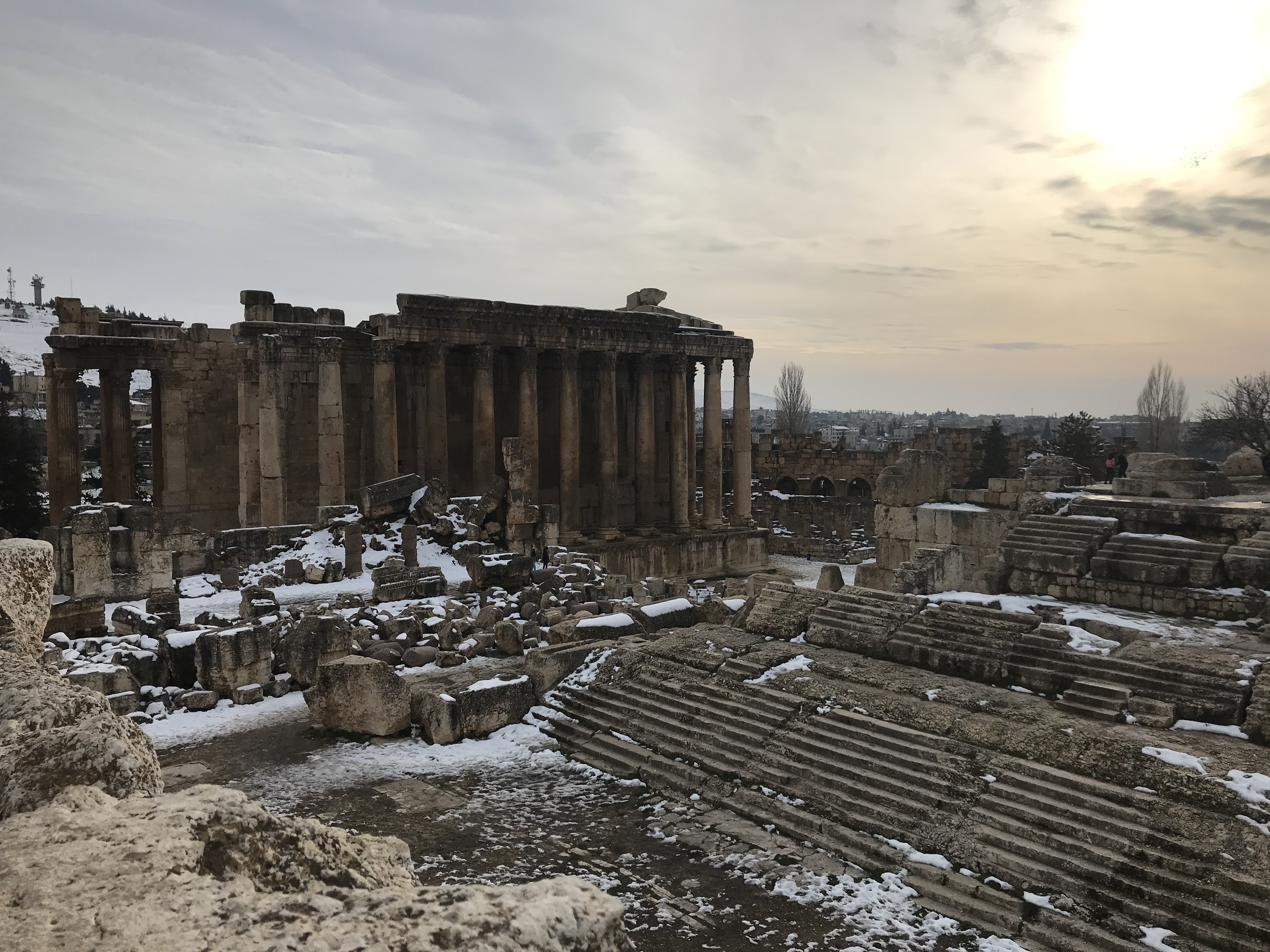 Landscape scene of Roman ruins at Baalbek, with the ~20 meter tall temple on the right and various staircases and columns lying in ruins on the surrounding ground. There is a dusting of snow among the ruins, and the sky is hazy with the sun glowing brightly on the top right.