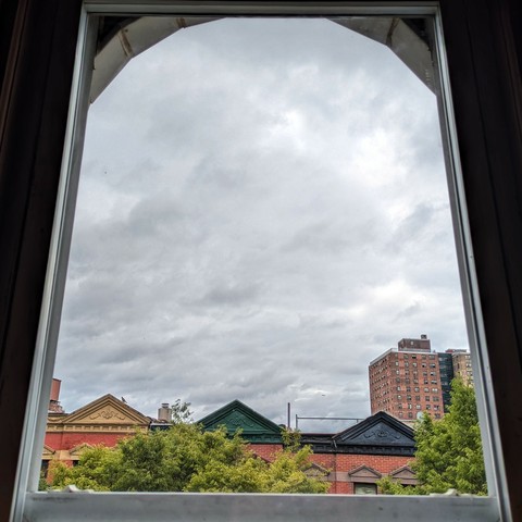 Looking through an arched window three hours after sunrise the sky is full of chunky grey rain clouds. Pointed roofs of Harlem brownstones with red brickwork are across the street, and a taller apartment building can be seen in the distance. The green tops of two trees are on the bottom and right.