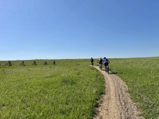A group of cyclists rides on a dirt path through a green field under a clear blue sky.